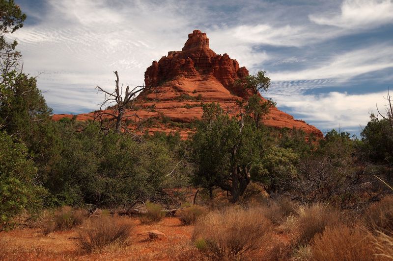 Bell Rock, a bell-shaped red sandstone butte, rising above juniper and dry grass under a streaked sky south of Sedona