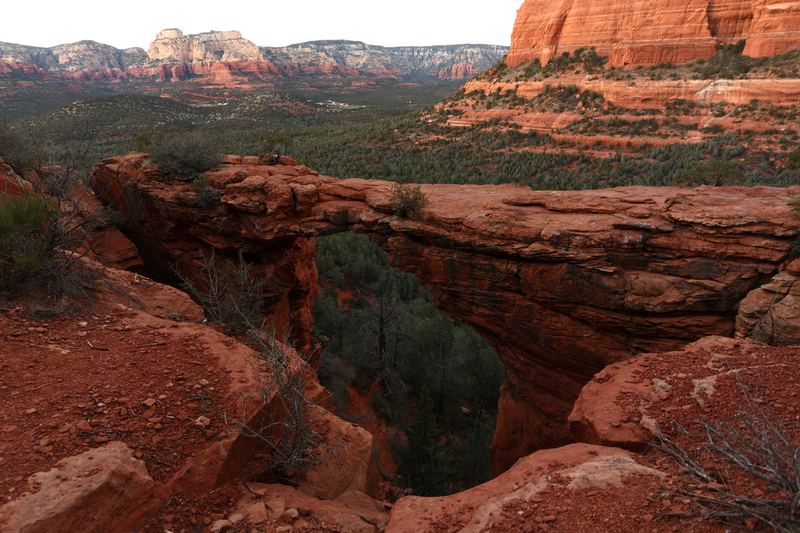 Devil's Bridge, a natural red sandstone arch, spanning a drop above juniper woodland in the Dry Creek basin, with Sedona's mesas behind it