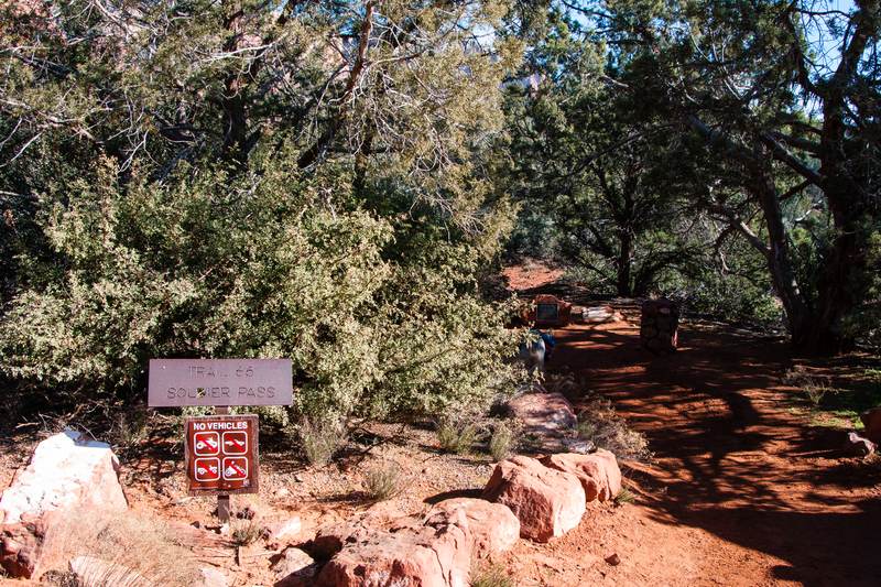 A Trail 66 Soldier Pass sign and a No Vehicles sign at the start of a red dirt trail through juniper and manzanita, Sedona